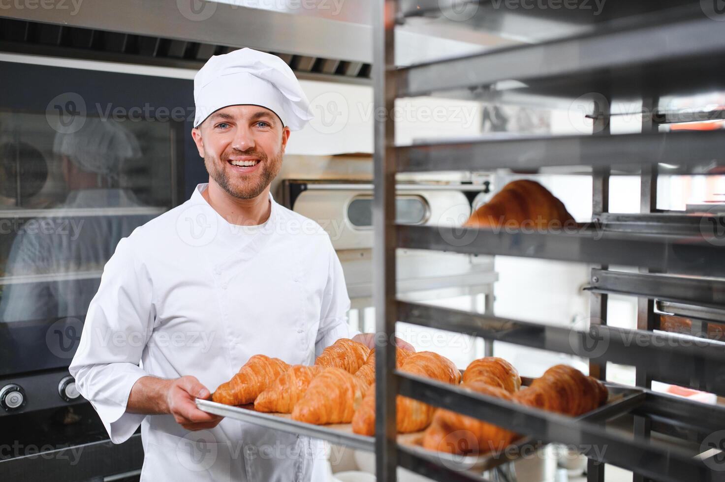 Smiling baker holding tray with fresh cooked croissants in bakehouse photo