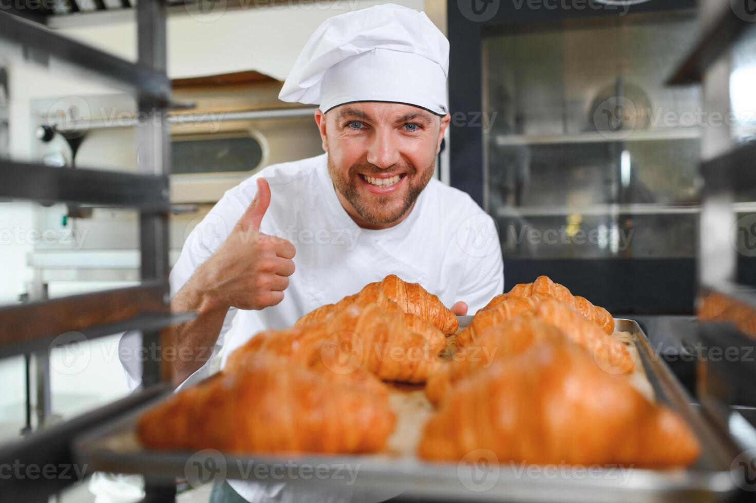 Handsome baker in uniform holding tray full of freshly baked croissants at the manufacturing photo