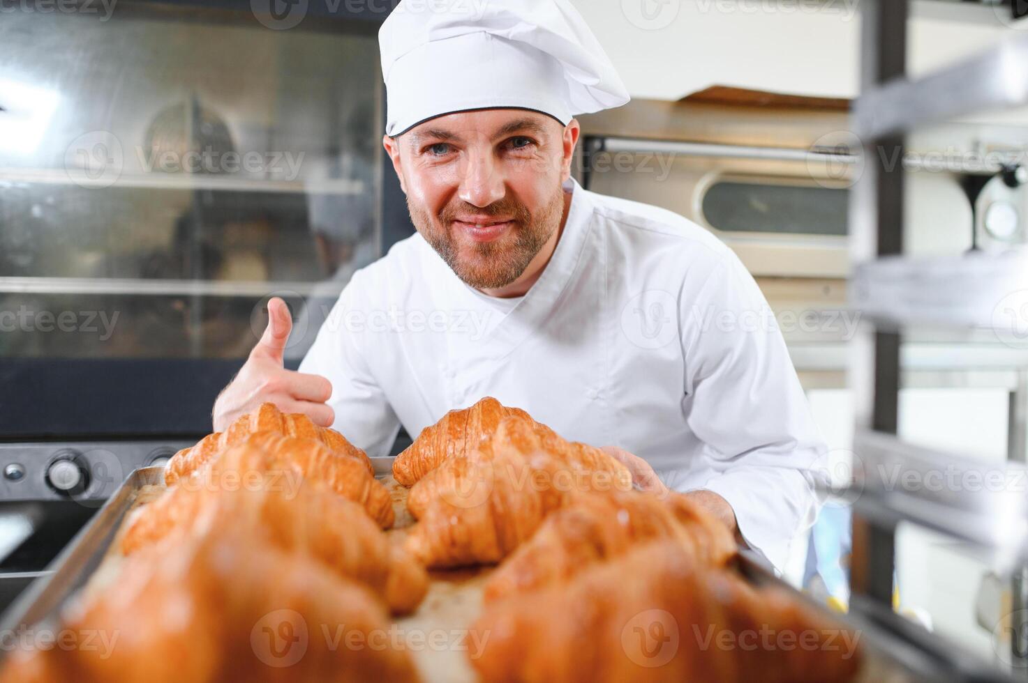 Smiling baker holding tray with fresh cooked croissants in bakehouse photo