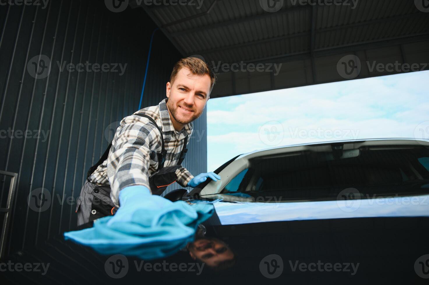 Car detailing, wash and cleaning concept. Young bearded male professional car wash worker in black rubber gloves, holding microfiber and wiping body of luxury car photo