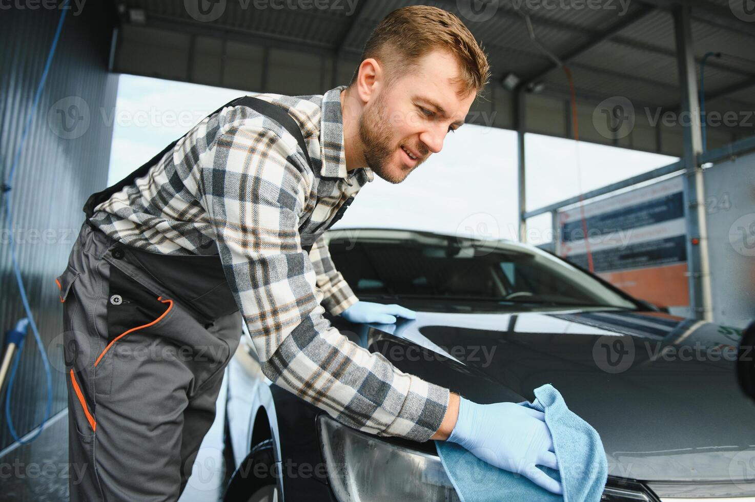 Bearded car wash worker man, wearing protective clothes uniform overalls, cleaning modern car with microfiber cloth photo