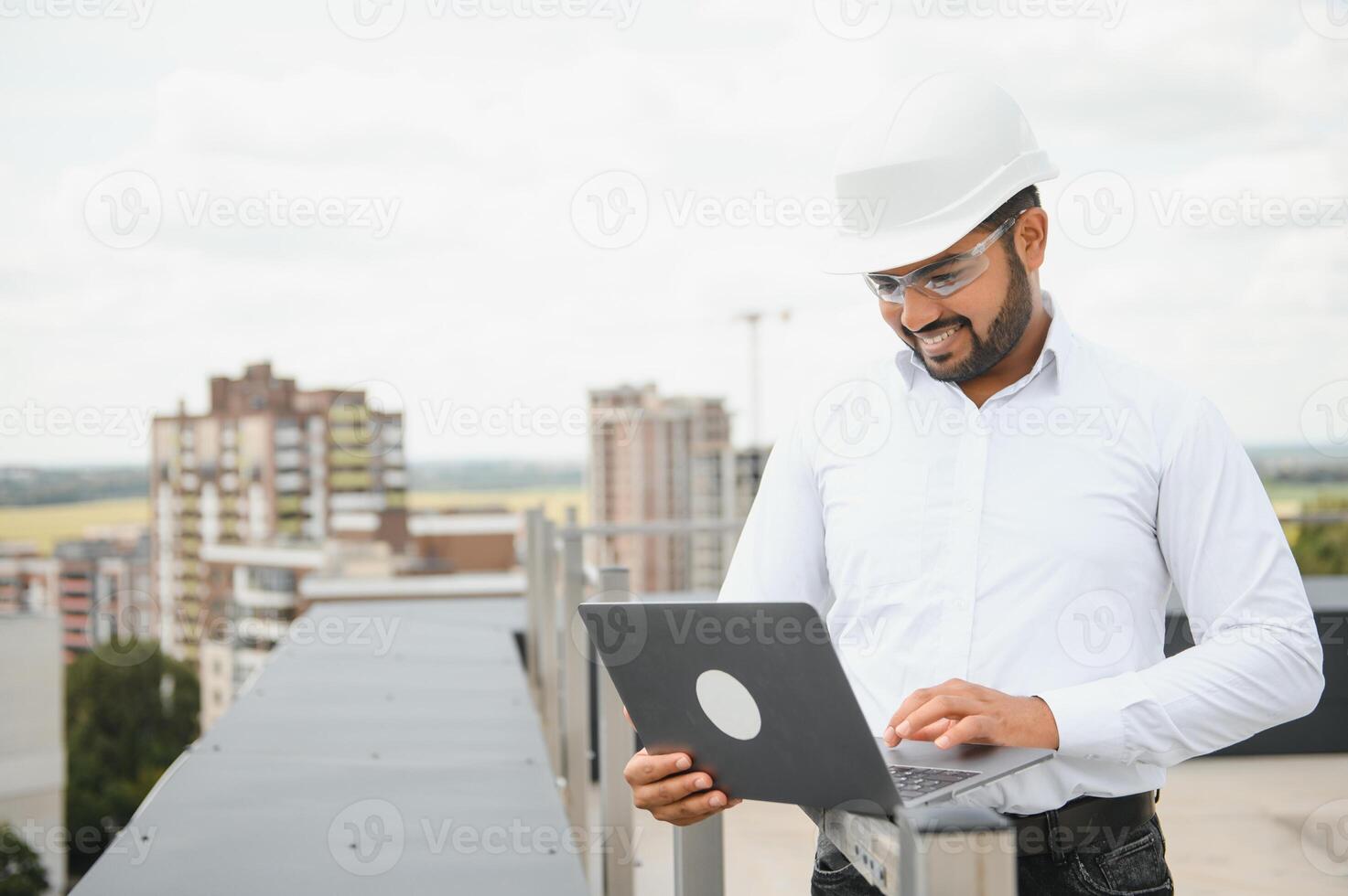 Asian Indian male contractor engineer with hard hat standing in front construction site, inspecting the progress photo