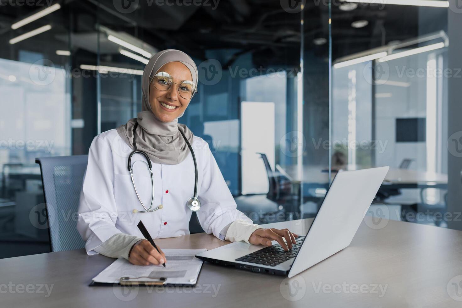 Portrait of female doctor in hijab, muslim working in modern office inside clinic, smiling and looking at camera, filling documents, using laptop photo
