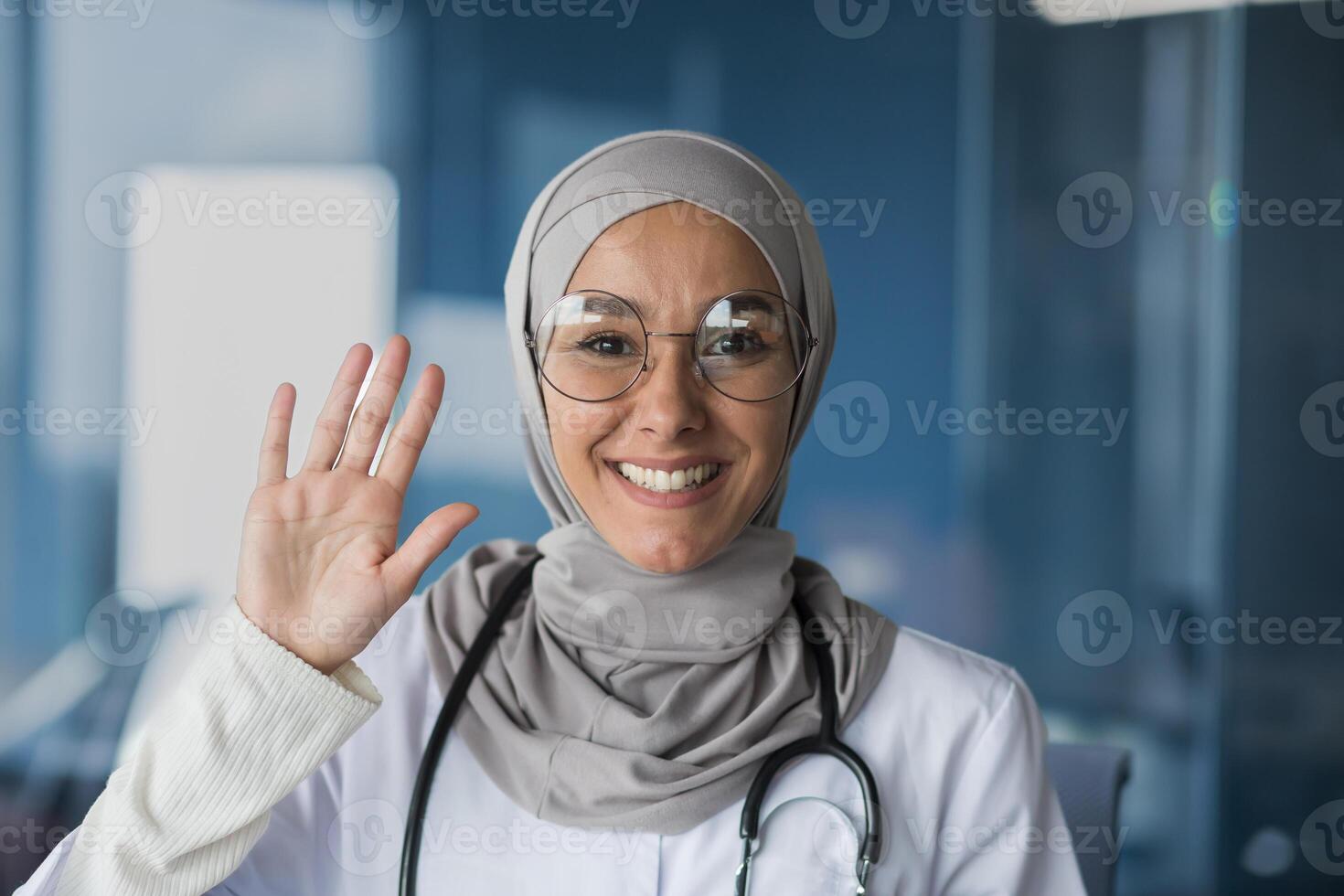 Portrait of a young beautiful woman, a Muslim, Arab woman doctor. She stands dressed in a hijab, glasses and with a stethoscope, looks and waves at the camera, smiles, greets. photo
