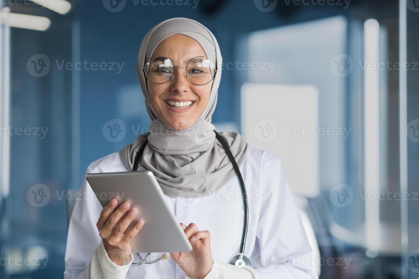 Portrait of a Muslim female doctor in a hijab with a tablet in her hands, the doctor works in the office of a modern clinic, smiles and looks at the camera. photo
