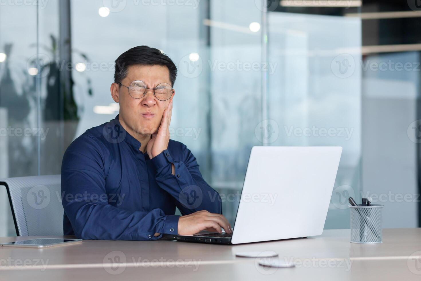 Asian sick in office, businessman has a toothache, man sitting in office inside building working at work using laptop, upset boss in shirt near window. photo