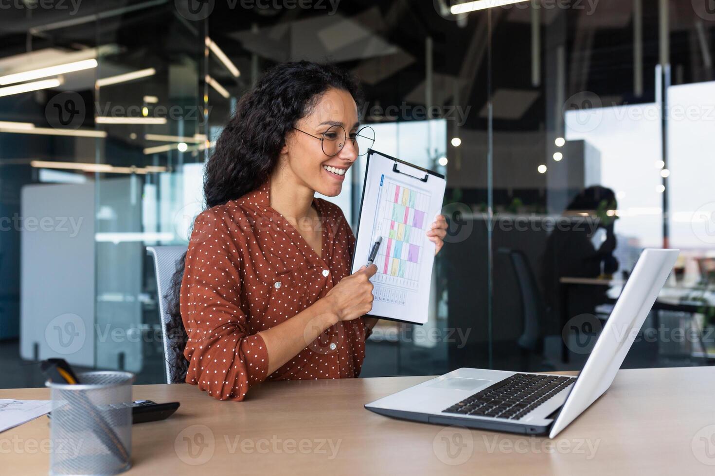 Business woman explains report indicators and financial results on call, Hispanic woman smiles and looks at laptop web camera, female worker works inside modern office building with reports. photo