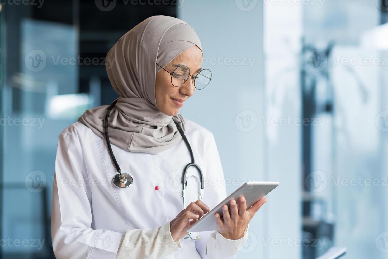 Close-up photo. A young Arab woman, a Muslim doctor is standing in the hospital in the office in a hijab and with a stethoscope. She holds a tablet in her hands, concentrates on working, typing. photo