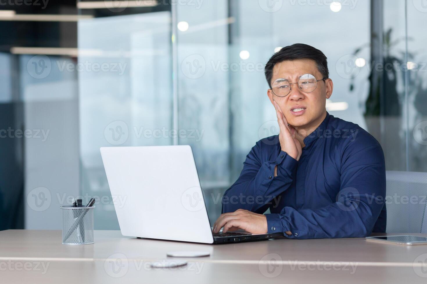 Asian sick in office, businessman has a toothache, man sitting in office inside building working at work using laptop, upset boss in shirt near window. photo