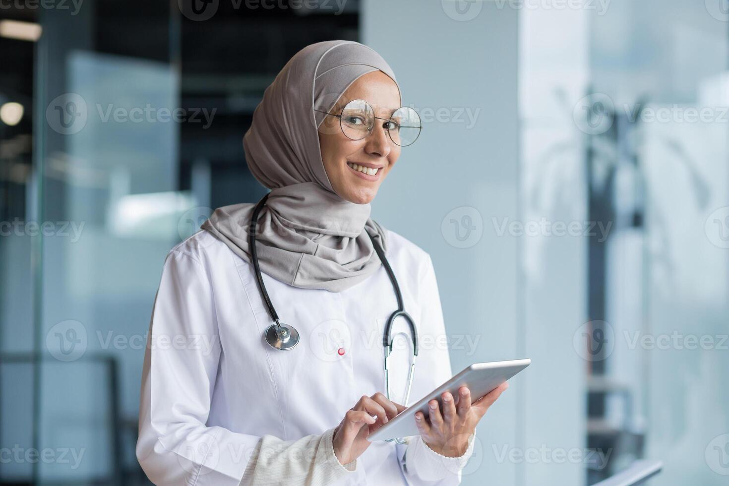 Portrait of a Muslim female doctor in a hijab with a tablet in her hands, the doctor works in the office of a modern clinic, smiles and looks at the camera. photo