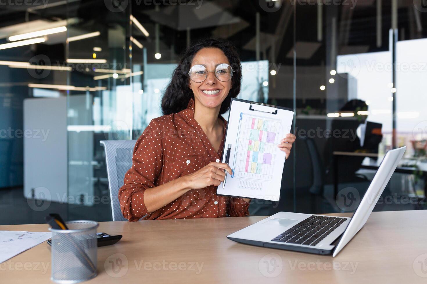 Portrait of happy and smiling woman accountant financier, Hispanic woman looking at camera and smiling showing document with financial indicators graphs, businesswoman working inside office. photo