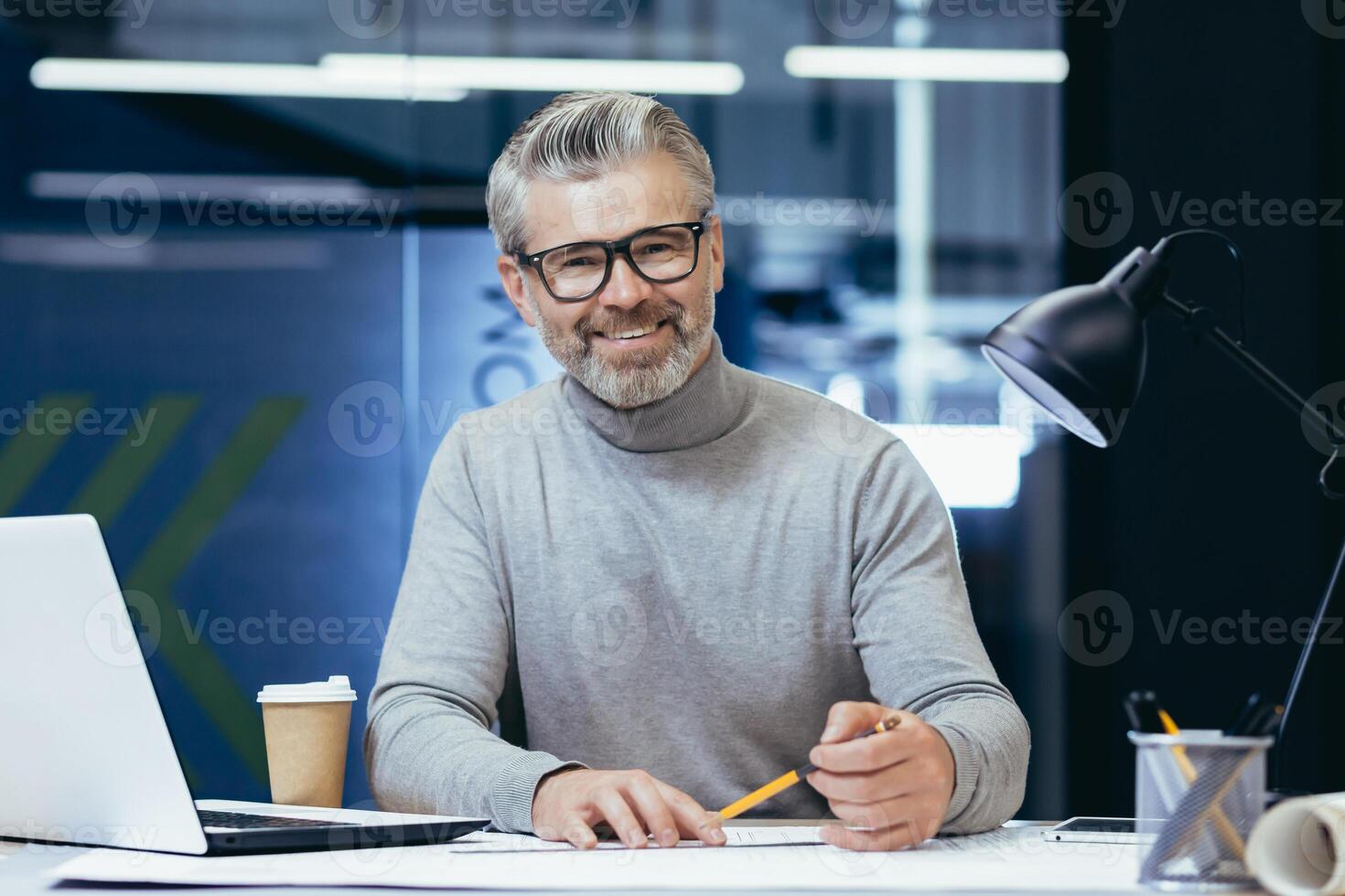 Portrait of an older gray-haired male teacher. He sits at a desk with a laptop, looks at the camera, smiles, holds a pencil in his hands. Teaches online, remotely. photo