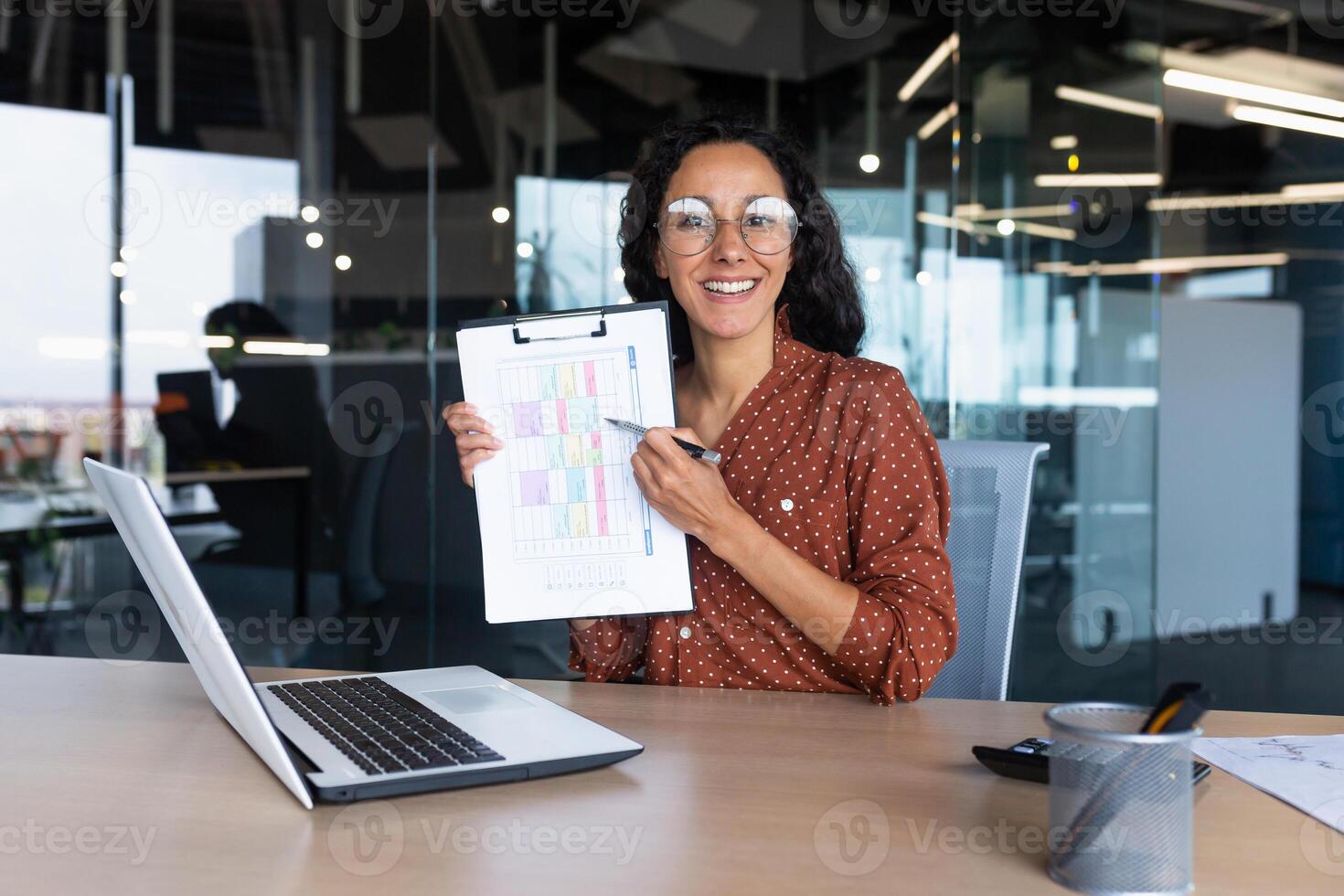 Portrait Hispanic business coach, woman smiling and looking at camera, showing documents with economic indicators to camera, teacher teaching remotely, mentor using laptop for online learning. photo