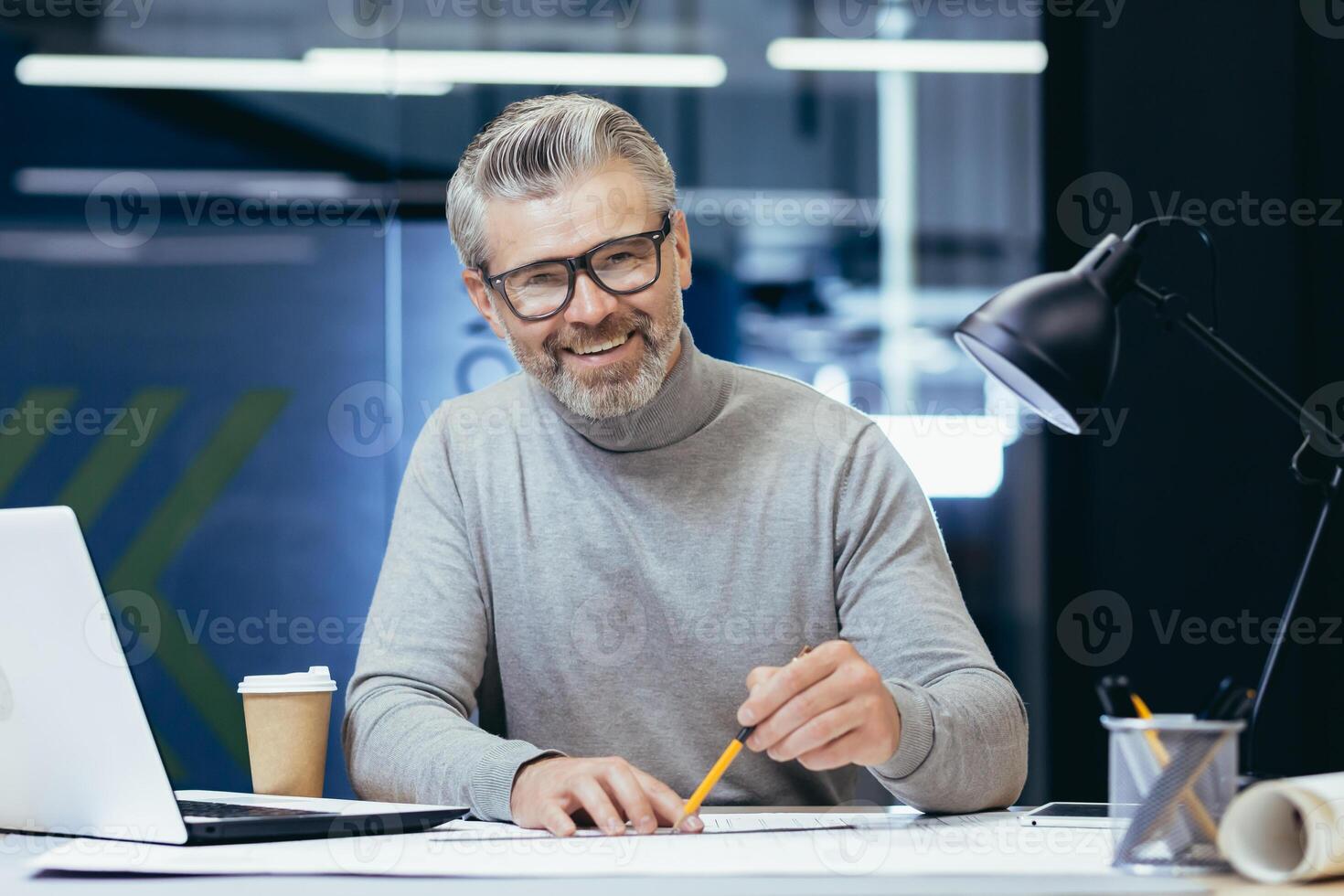 Portrait of senior architect, gray haired designer smiling and looking at camera, man mock up plan use laptop and tablet computer at work inside studio office. photo