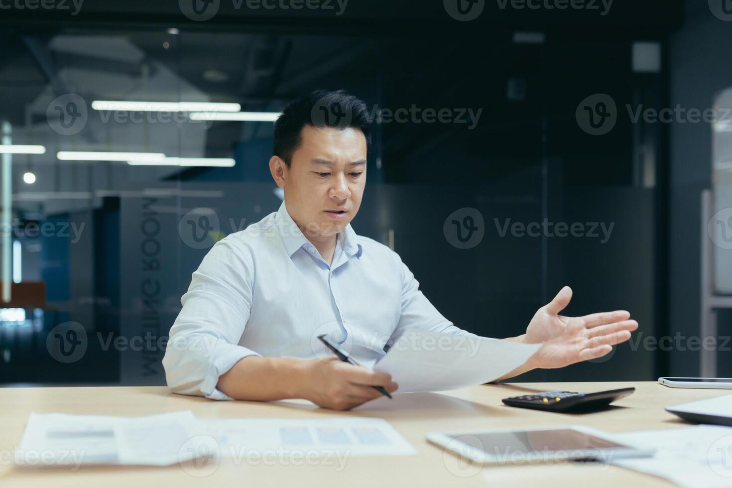 Bankruptcy. Financial problems. A serious asian man sits in the office at a table with a laptop, holds documents, bills, an agreement in his hands. He looks worriedly at the papers, spreads his hands. photo