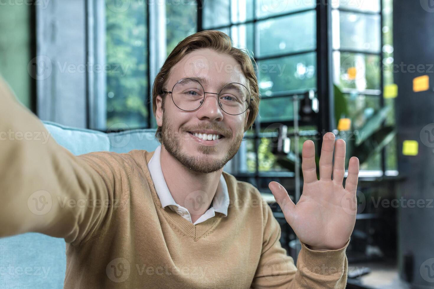 Man working in modern office, happy blond businessman smiling and looking at smartphone camera, holding hand up greeting gesture, worker in glasses taking selfie, call with friends photo