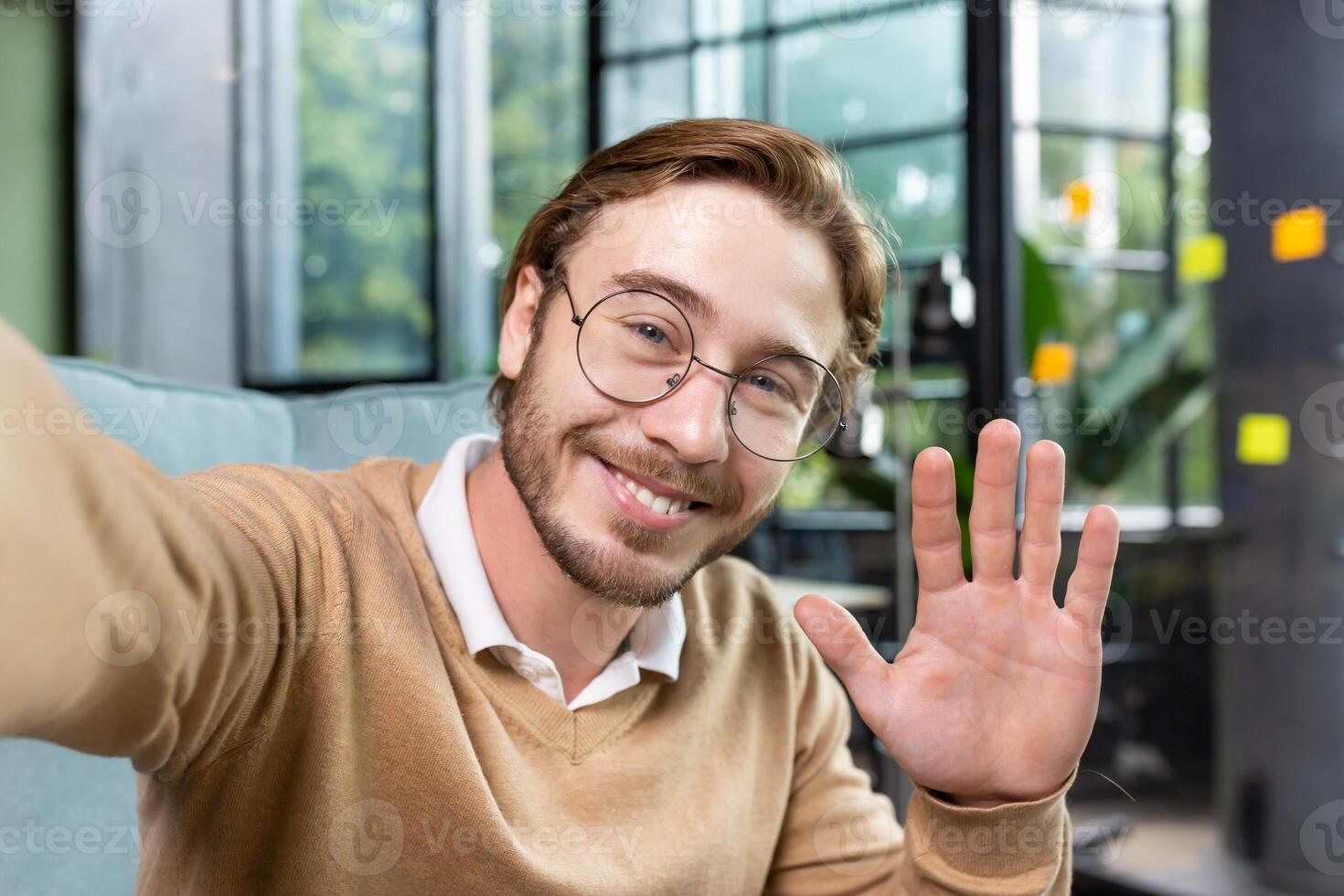 Close-up photo. Young male blogger, student, businessman waving, greeting and talking to camera. Sitting at the desk in the office, smiling and taking pictures. photo