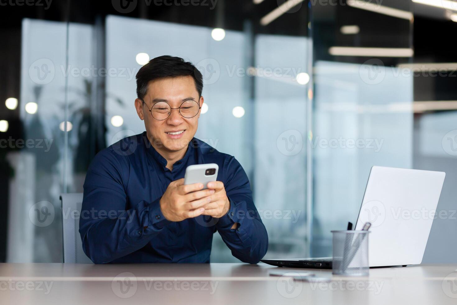 Cheerful and smiling asian businessman inside office using phone, man typing message and browsing online pages and watching , boss in glasses and shirt at work. photo