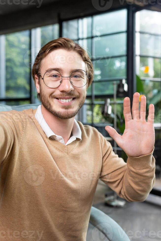 Vertical photo. Close-up photo. Young man freelancer, blogger greets and talks to the camera, smiles, waves, records , blog. photo