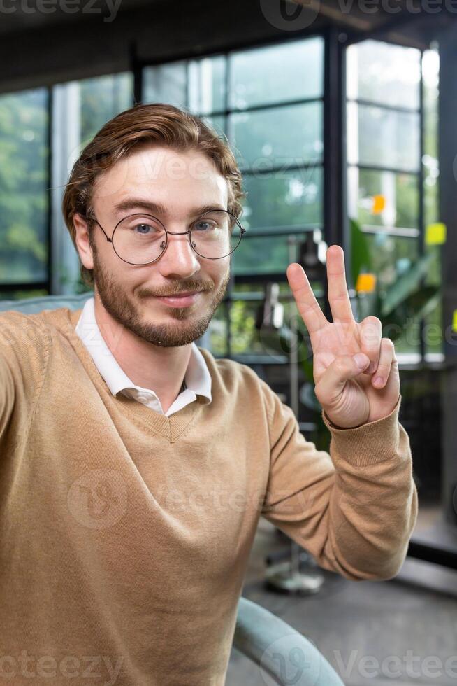 Man in office wearing shirt and glasses smiling and looking at smartphone camera, businessman taking selfie and talking on call using smartphone, programmer waving hand greeting gesture photo