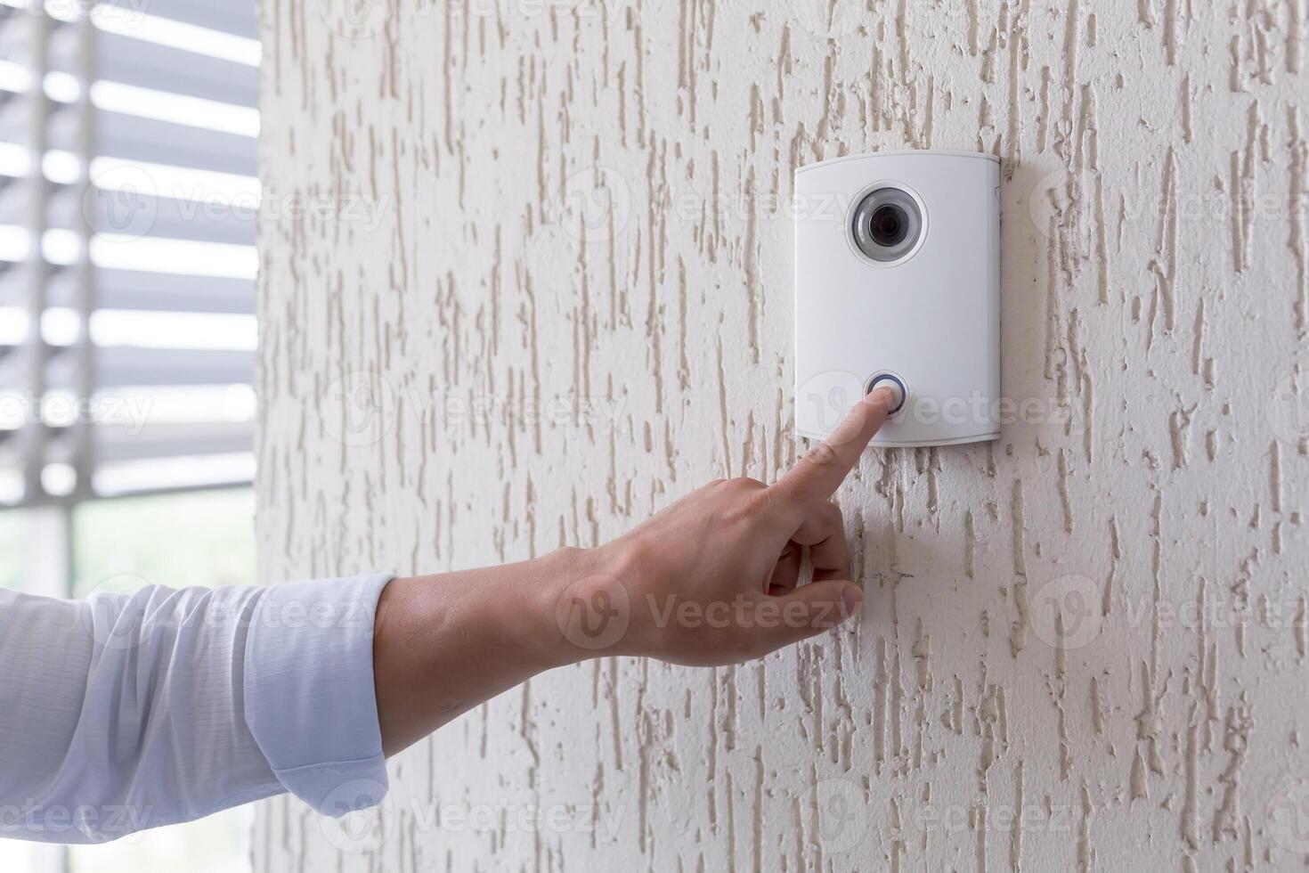 Hand of a man in a white shirt pressing the button of a door knocker on a white wall of a house, a smart door knocker with a camera for communication photo