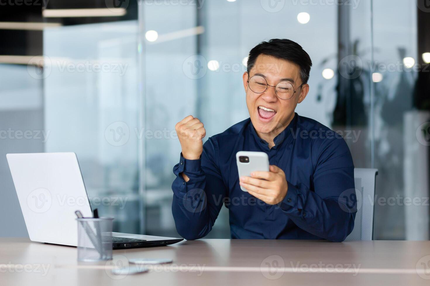 A happy young man, an Asian businessman, a student sits in the office at a table with a laptop, uses a mobile phone. Rejoices, celebrates, received good news, win, shows with hand yes photo