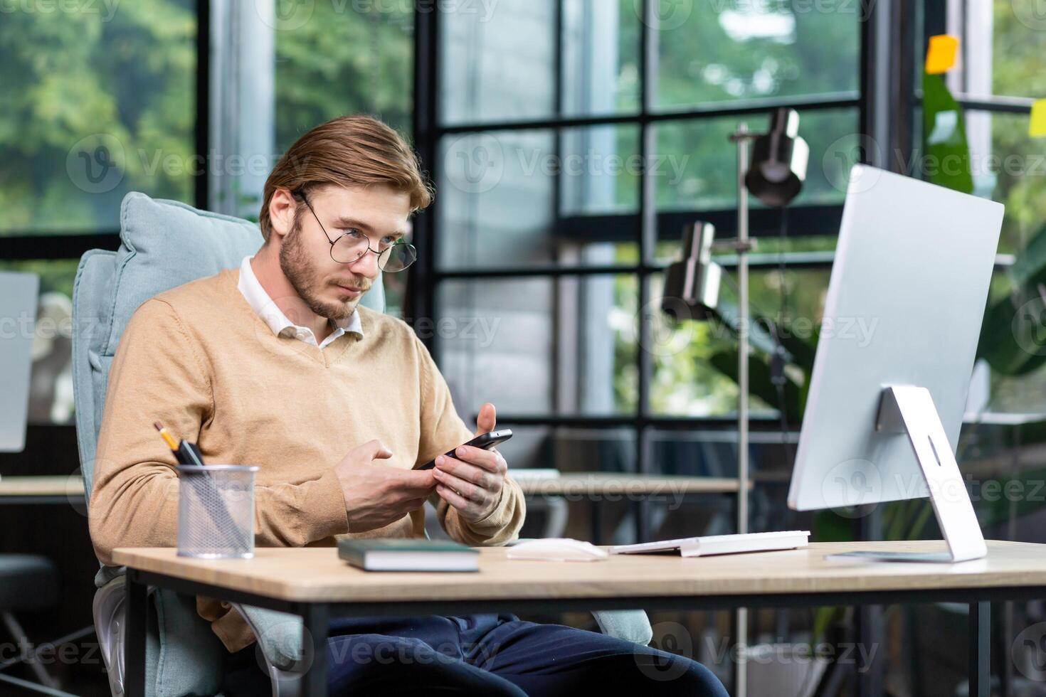 A young programmer, freelancer sits at the desk in the office, works at the computer. Holds a phone in his hands, uses a gadget. Writes a message, checks mail. photo
