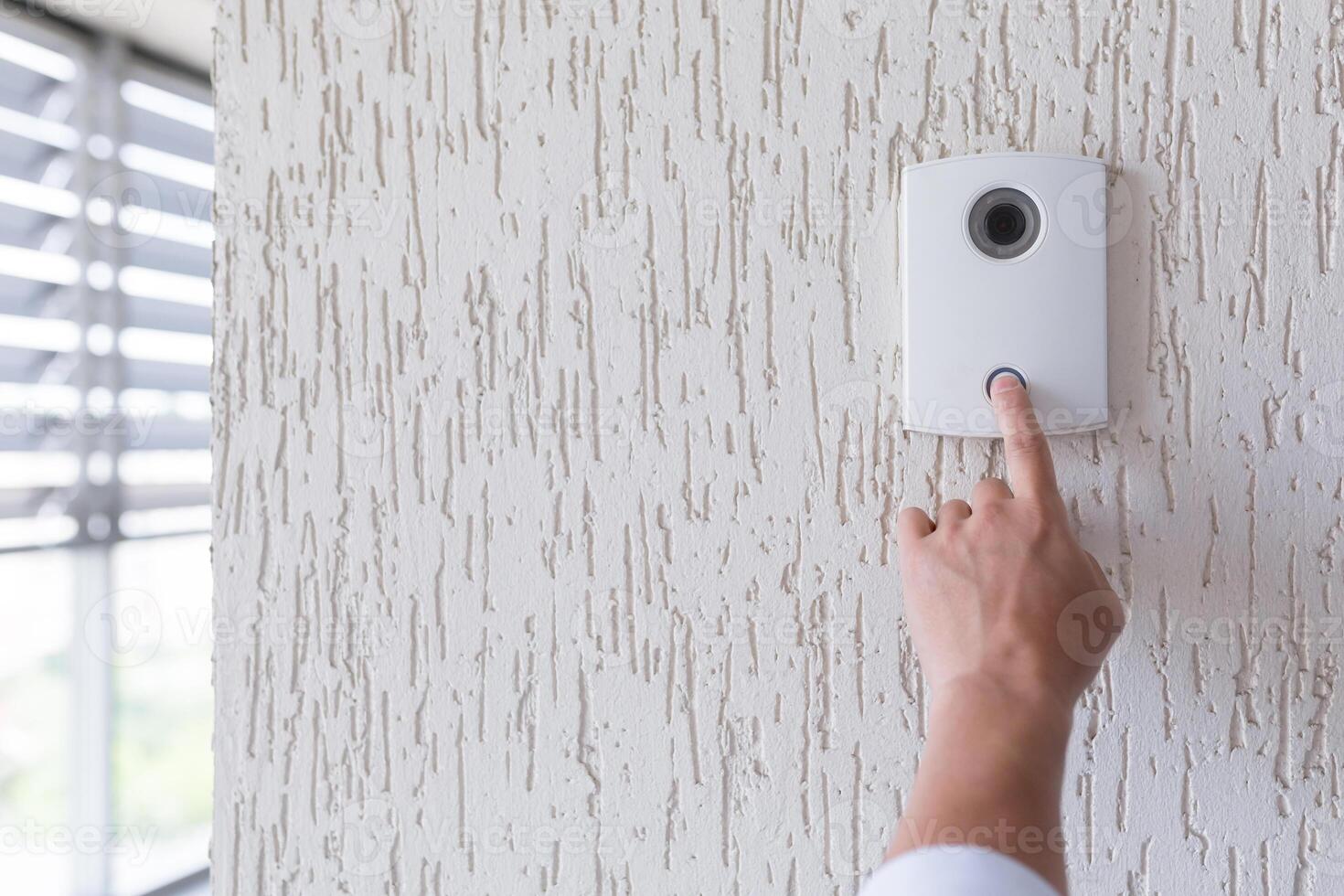 Hand of a man in a white shirt pressing the button of a door knocker on a white wall of a house, a smart door knocker with a camera for communication photo