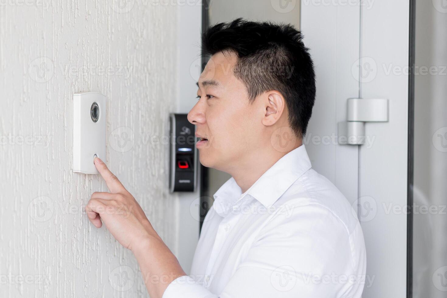 An Asian man stands at the door of a house, office, hotel and presses the door bell of an automatic door with his finger. Speaks through loudspeaker, intercom, camera. photo