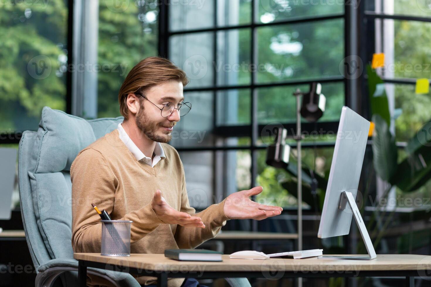 Portrait of a smiling young man sitting at the desk in the office in front of the computer and talking via call, conducting business training, online conference, webinar, business meeting. photo