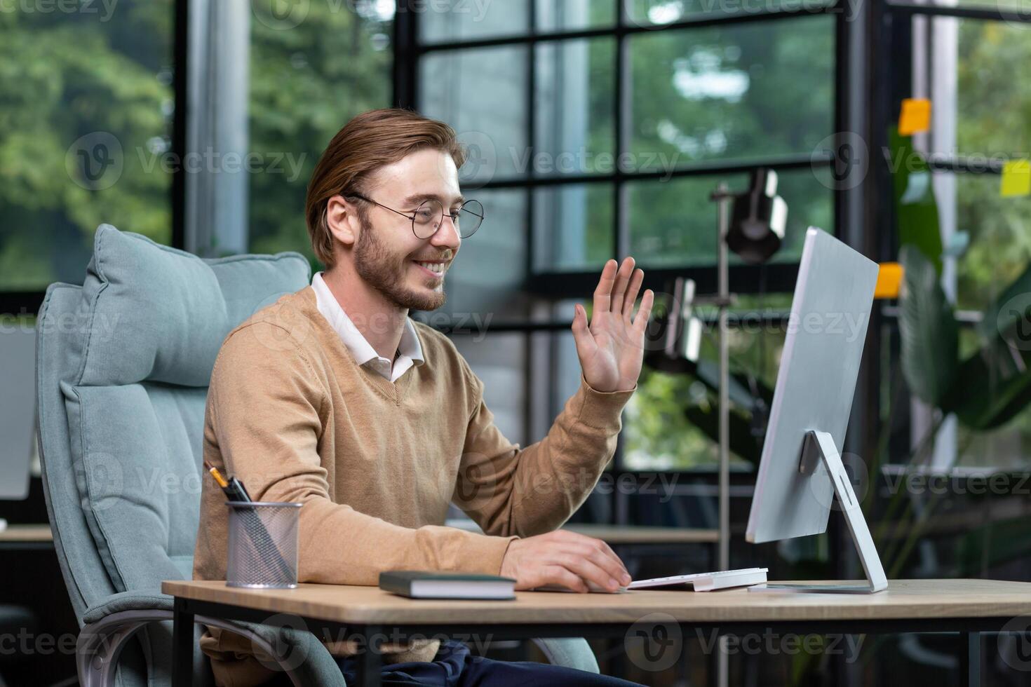 Successful, satisfied young boss conducts an online call with colleagues, sits at the table, looks at the computer monitor, waves at the webcam in greeting. photo
