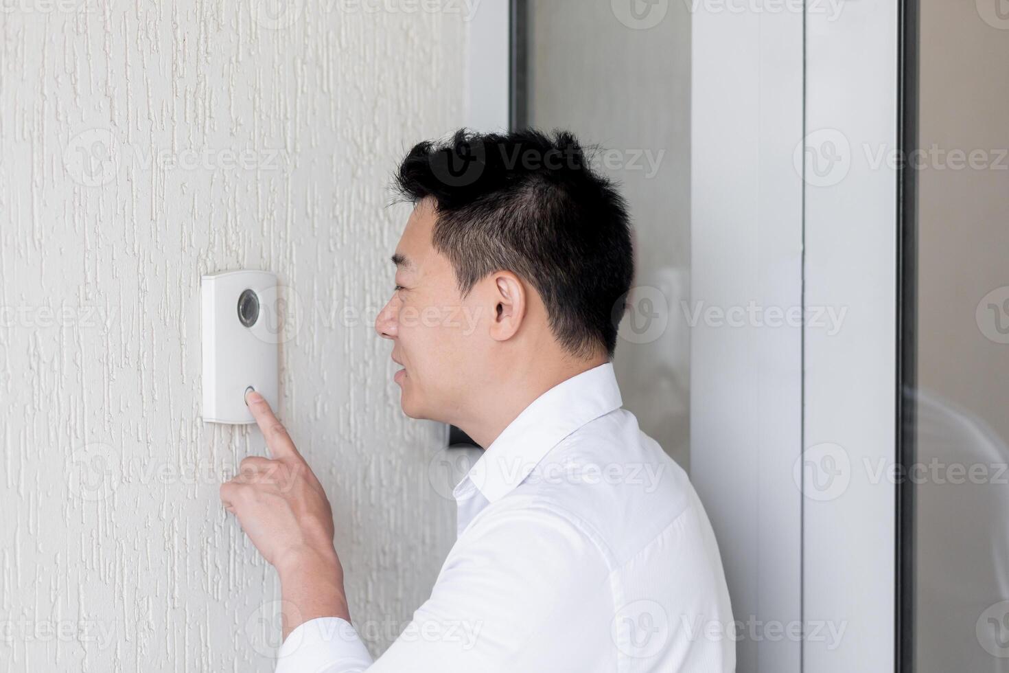 Close-up photo. A young Asian man calls with his finger on the electronic doorbell on the wall of the house, office. Speaks into the intercom photo