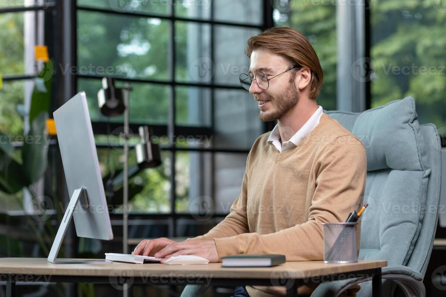 A young male programmer, freelancer sits in the office at the table near the window and works at the computer, typing on the keyboard. Smiling, wearing glasses and a beige sweater. photo
