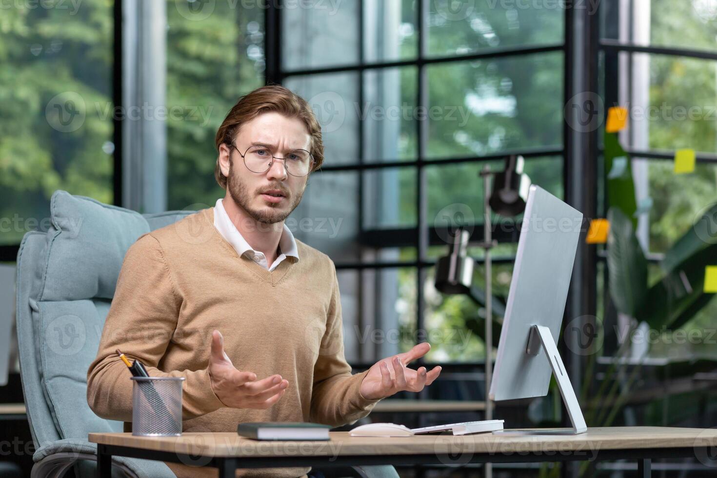 Solving a problem, making a decision. Portrait of a young freelancer, a programmer who looks worriedly and angrily at the camera, spreads his hands. Sitting in the office at the desk. photo