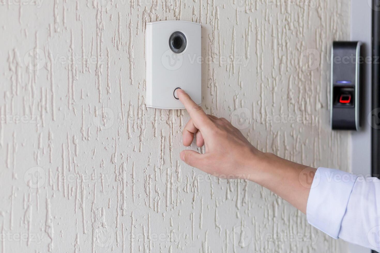 Hand of a man in a white shirt pressing the button of a door knocker on a white wall of a house, a smart door knocker with a camera for communication photo