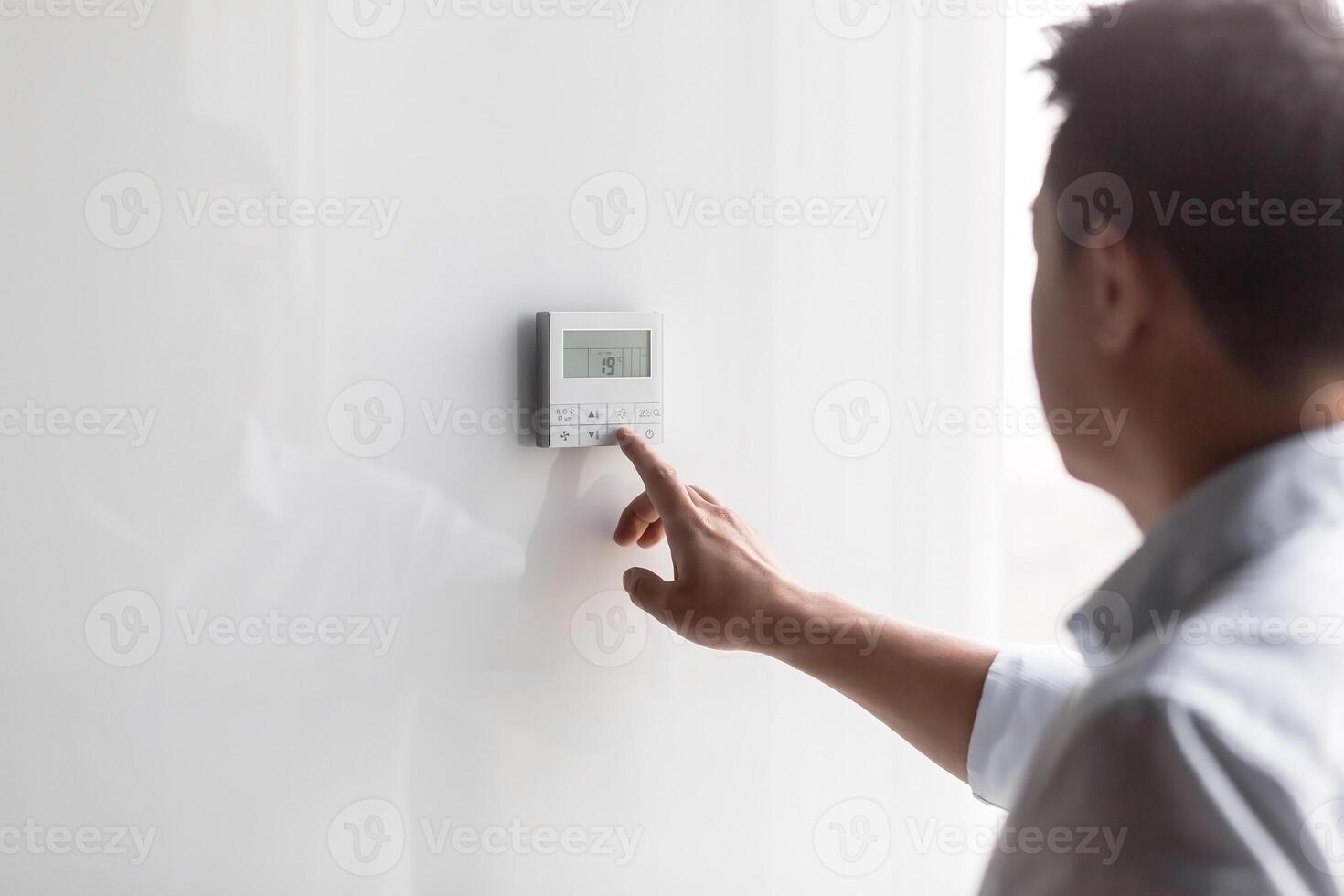 Close-up photo. The hand of a young man in a white shirt turns on the control buttons of the air conditioner hanging on the wall. Standing on the right photo