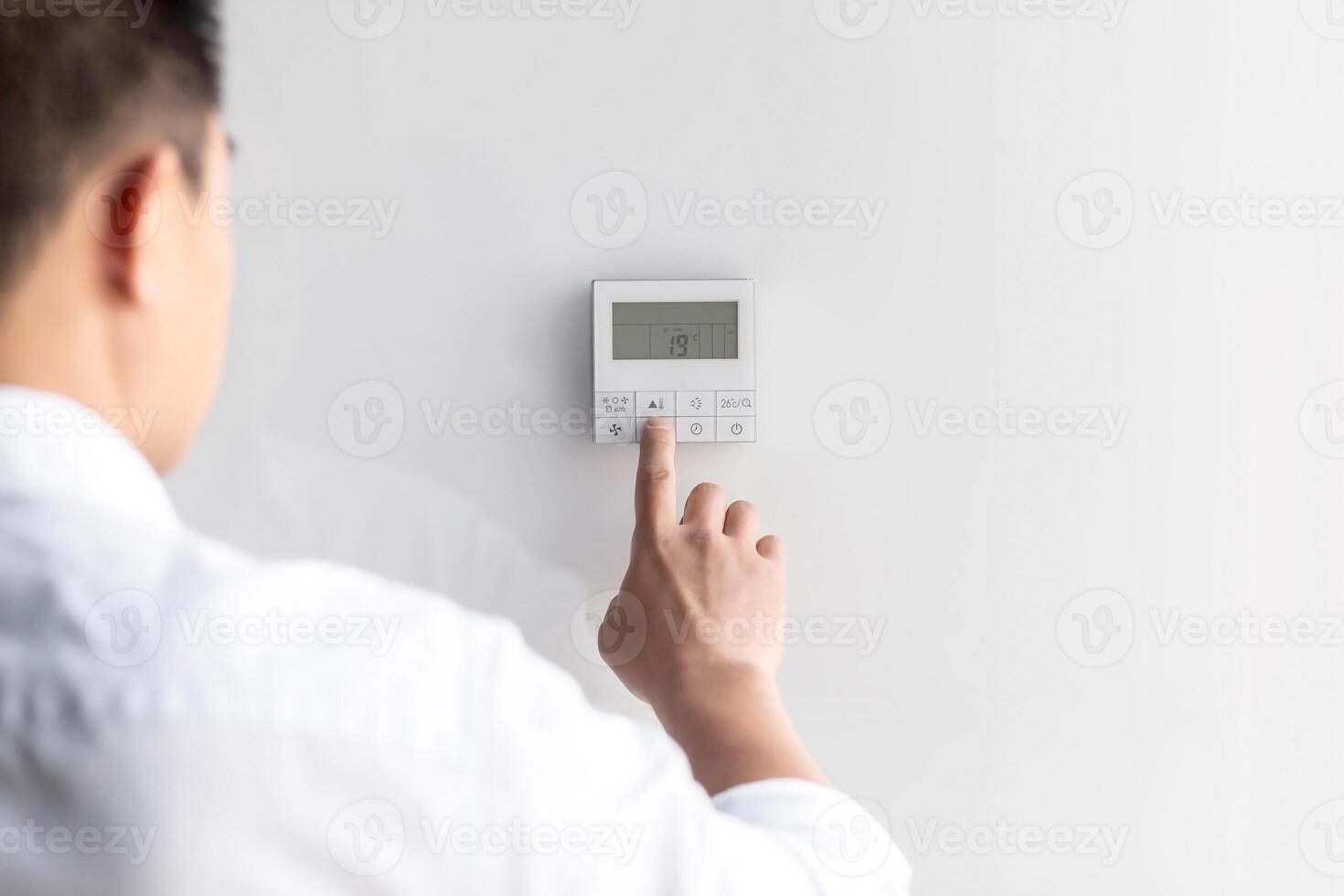 Close-up photo. The hand of a young man in a white shirt turns on the control buttons of the air conditioner hanging on the wall. Standing on the left photo