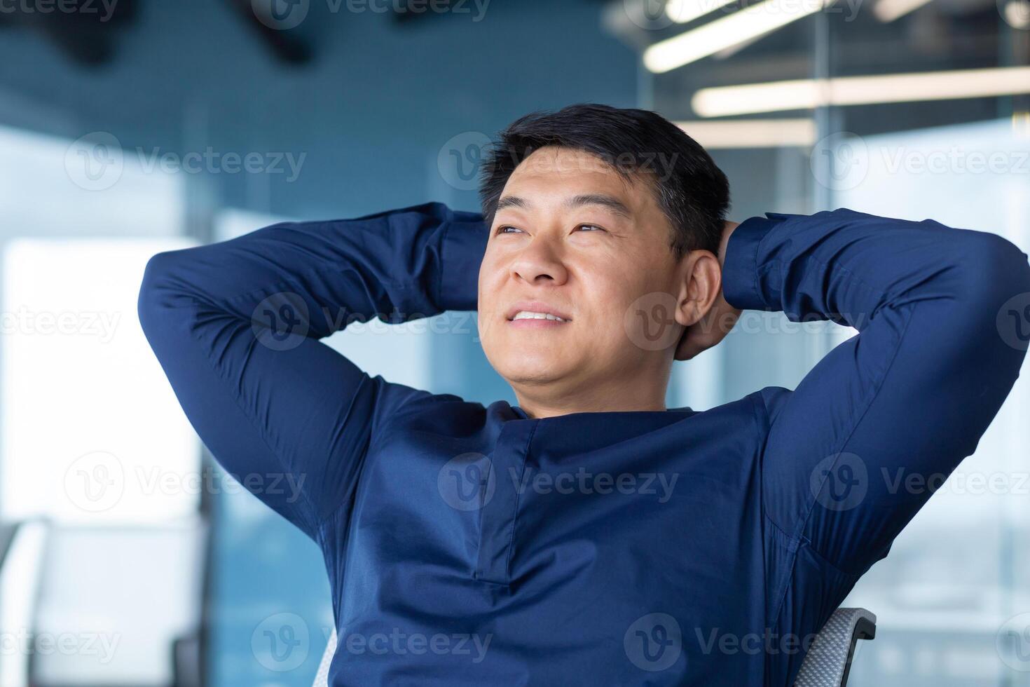 Happy businessman resting after successfully completed work asian man smiling and happy looking window thinking and dreaming about future project, man in shirt with hands behind head inside office. photo