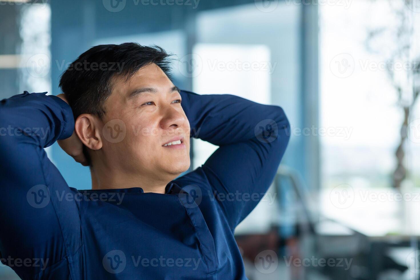 Asian man at home by the window resting and daydreaming, man in a shirt with his hands behind his head looks to the side and smiles, thinks about future plans. photo