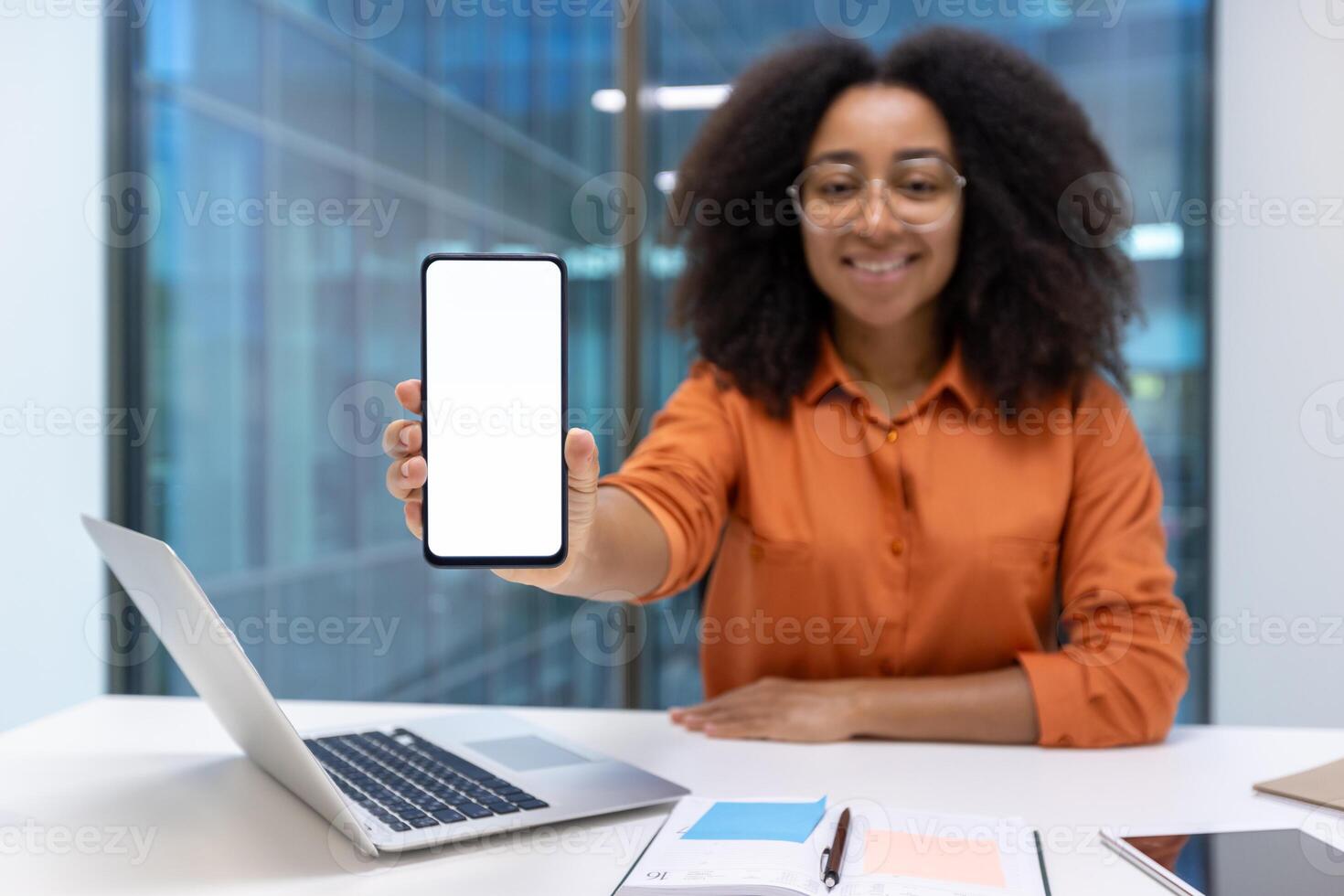 Young woman businesswoman displaying a smartphone at her desk in a modern office environment. She is wearing glasses and smiling, creating a professional yet approachable atmosphere. photo