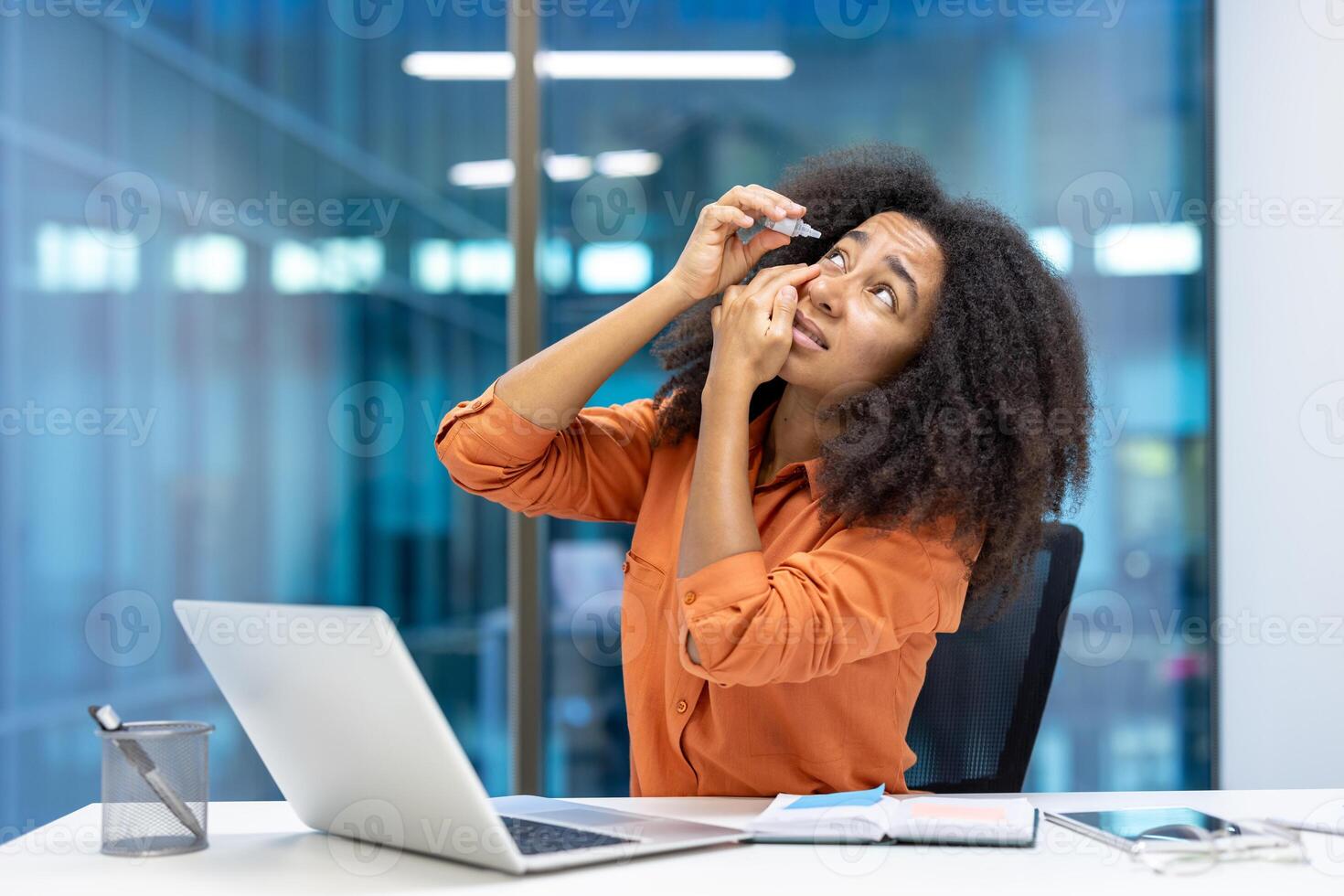 A woman sits at her office desk using eye drops. The workspace has a laptop, notepad, and a pen holder. She appears focused and attentive during her workday. photo