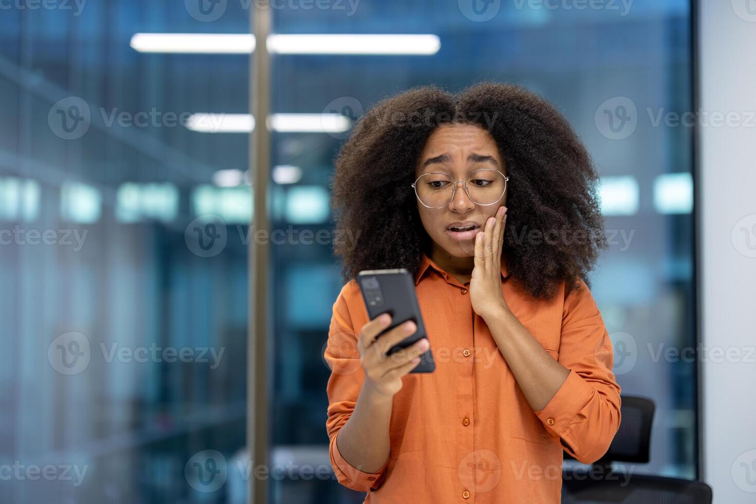 A young businesswoman appears shocked while checking something on her phone in a modern office setting. Her expression suggests receiving unexpected news or encountering a surprising situation. photo