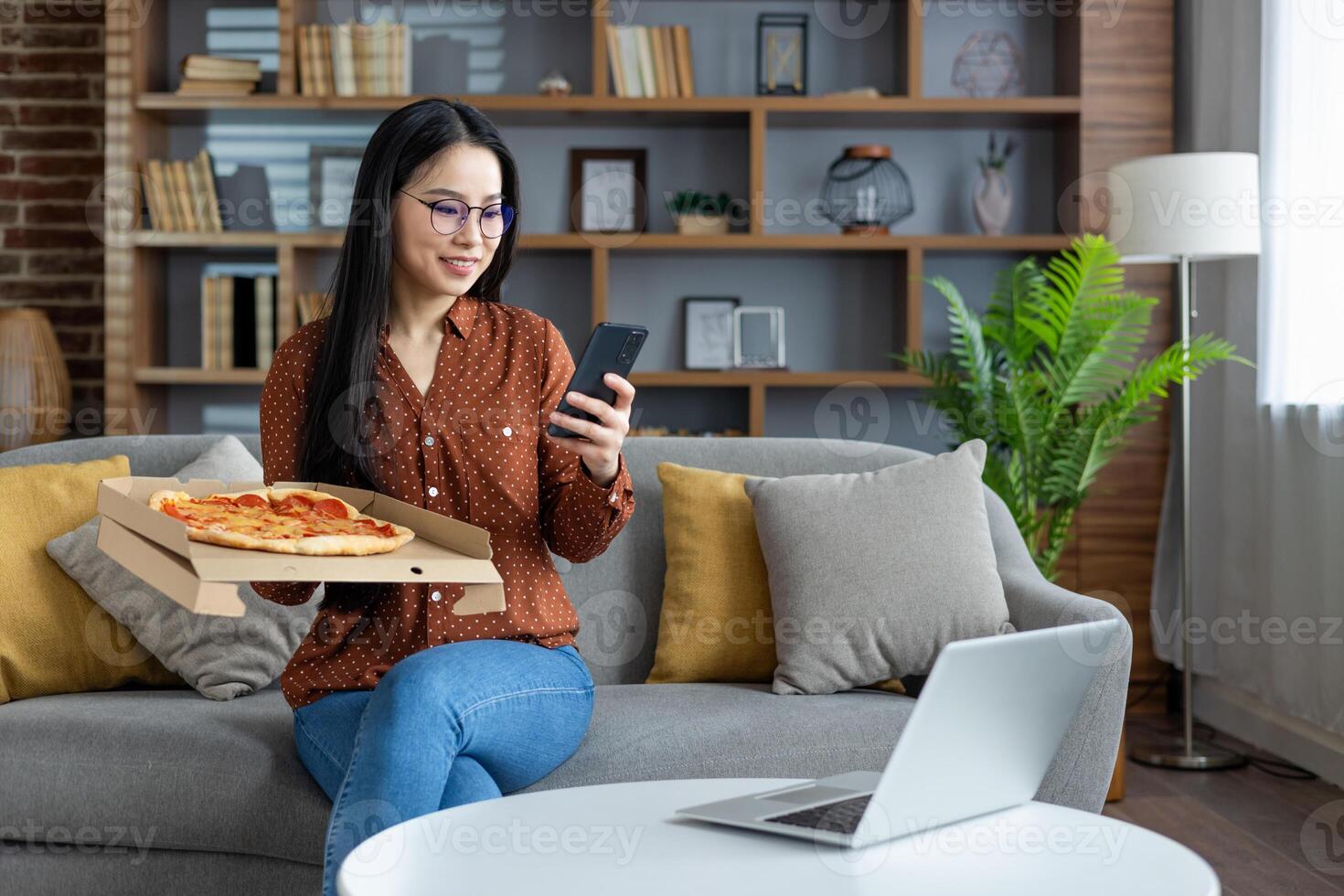 Asian woman enjoying pizza, using smartphone while sitting on sofa at home. Modern lifestyle embraces technology. Relaxed, casual moment with laptop, phone, and casual attire. photo