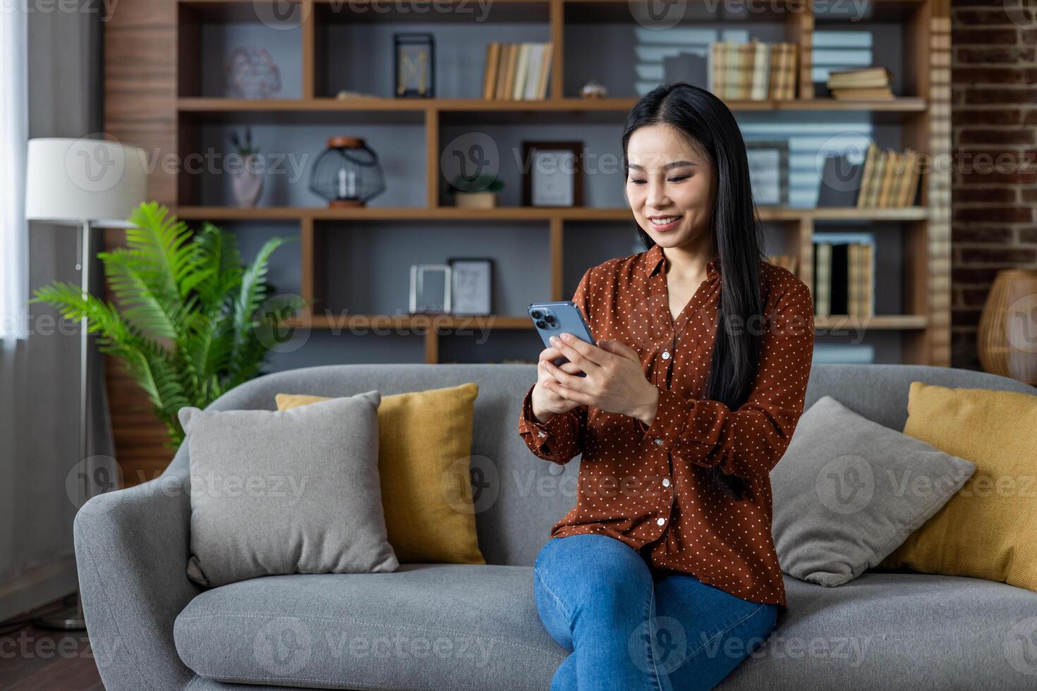Asian woman sits on sofa using smartphone, smiling while engaged in online activity. Modern home setting with stylish decor and comfortable seating creates relaxing environment. Technology in everyday life. photo