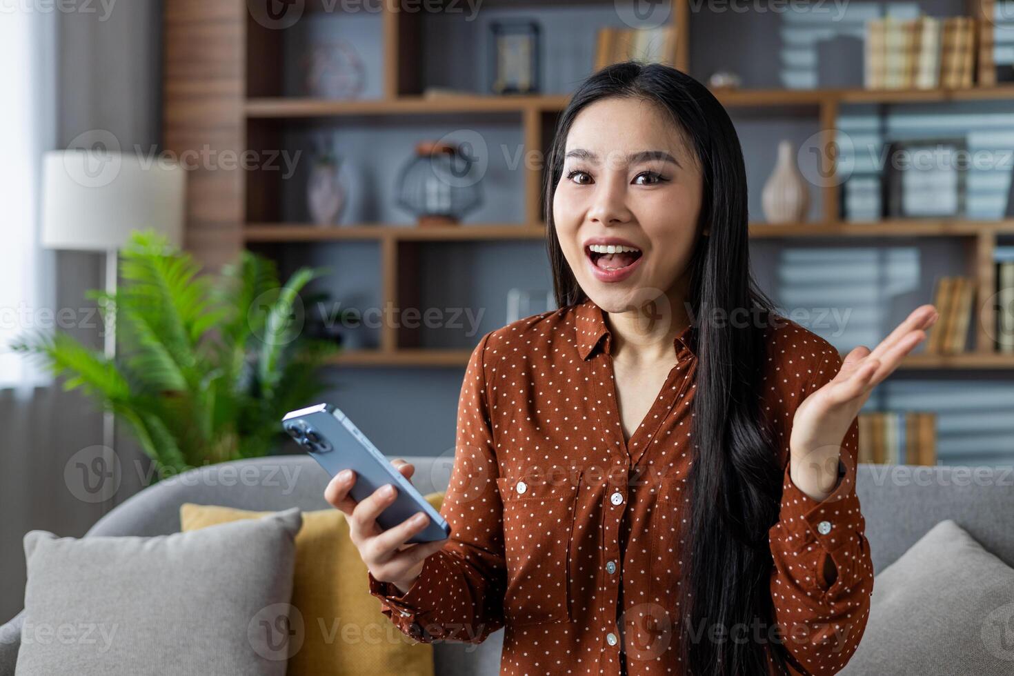 Asian woman delighted while holding phone at home. Exudes surprise and excitement in casual attire, against a bookshelf backdrop. Perfect depiction of modern lifestyle and joyful communication. photo