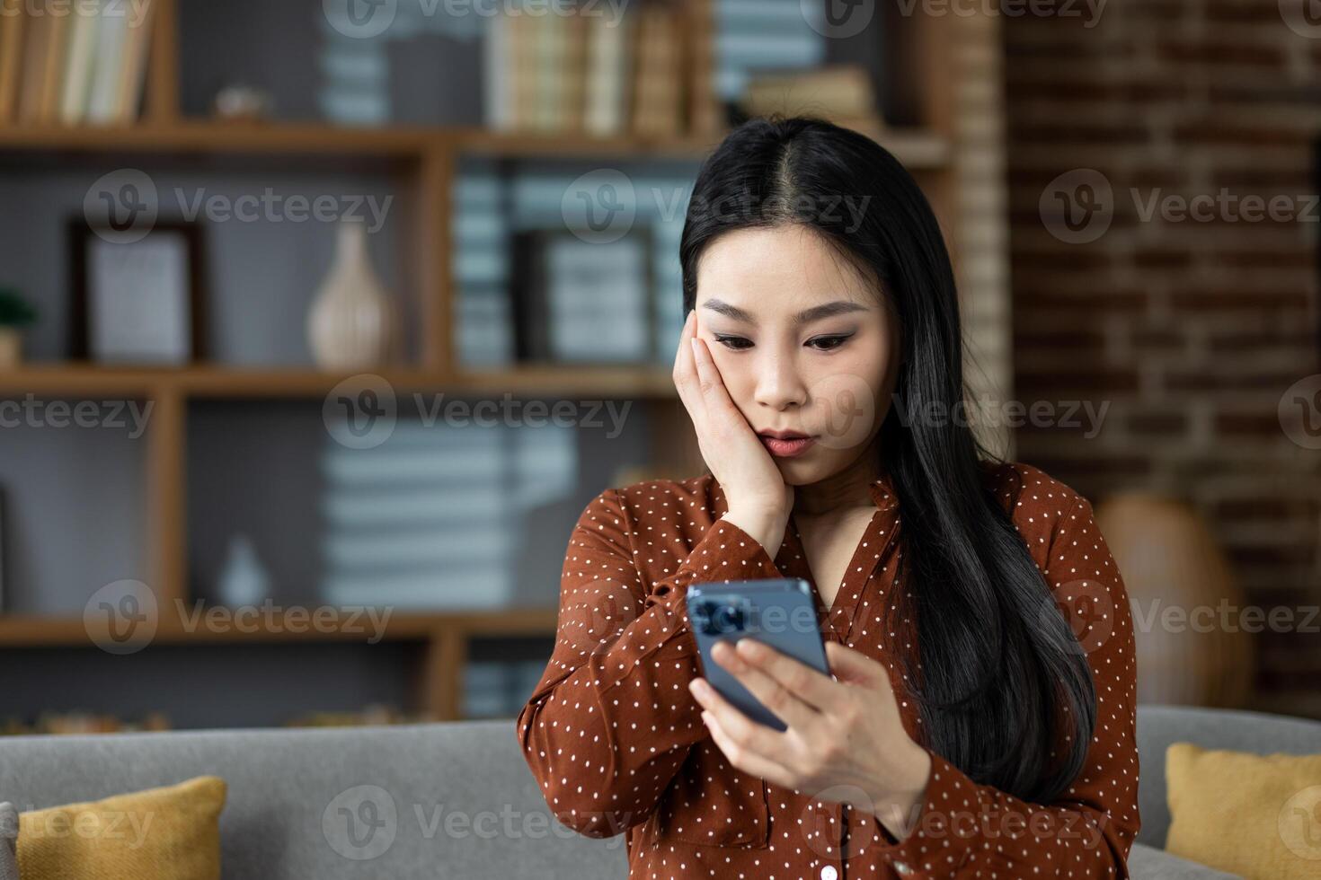 Asian woman at home looks surprised while holding smartphone. Modern lifestyle, home interior, and communication concept. Relaxed yet concerned expression. photo