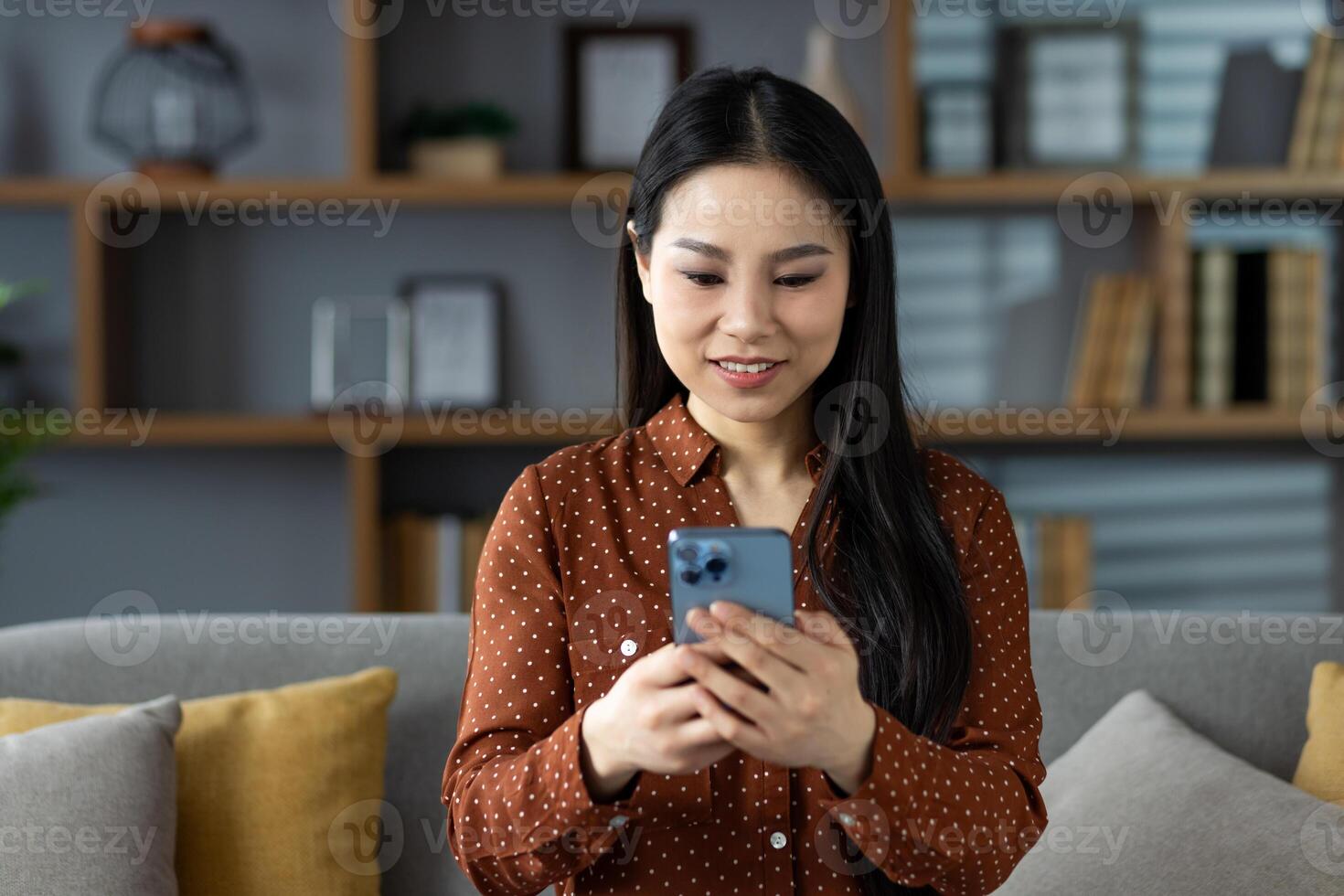 Asian woman casually dressed, sitting on couch at home, using smartphone. Smiling and engaged in activity, creating warm and relaxing domestic atmosphere. Perfect for lifestyle concepts. photo
