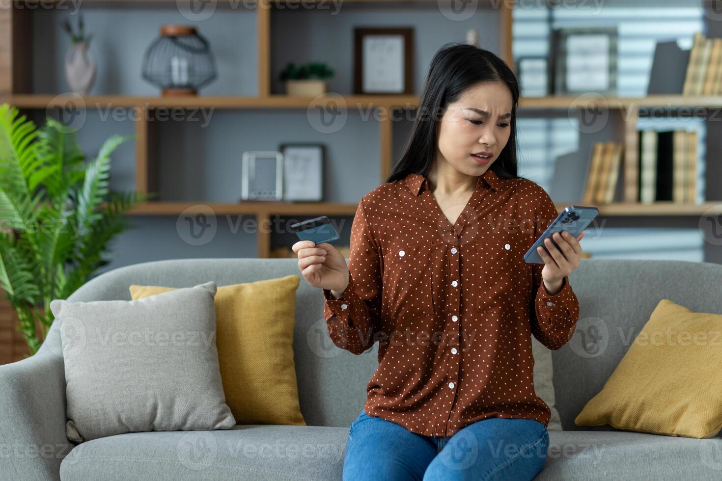 Concerned Asian woman using smartphone with credit card in hand sitting on couch. Concept of online shopping, banking issues, security concern, or financial stress. Focus on phone use. photo