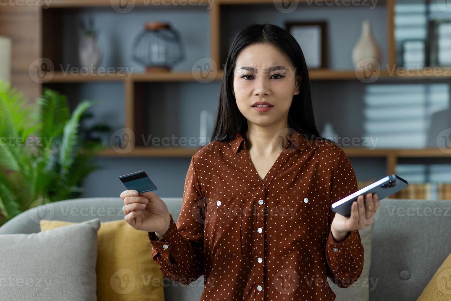 Asian woman puzzled, holding phone and credit card, expressing confusion about online shopping or mobile payment issues. Casual wear, cozy home setting, exploring technology use. photo