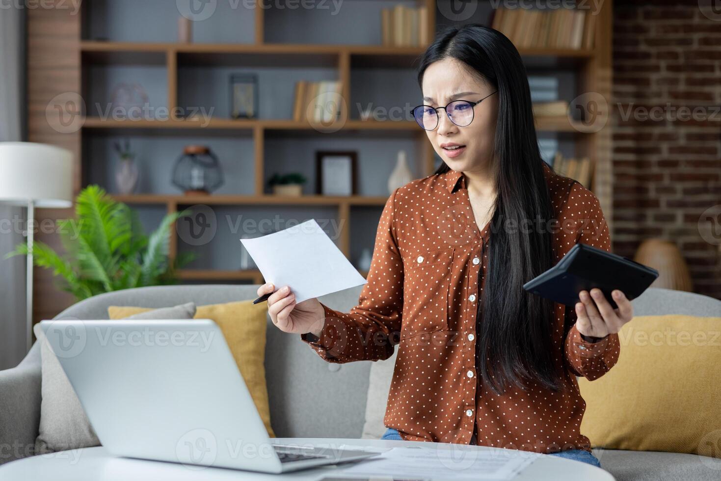 Asian woman managing finances, sitting on couch with laptop, calculator, examining bills, showcasing concern and focus. Represents budgeting, financial planning, home office environment. photo